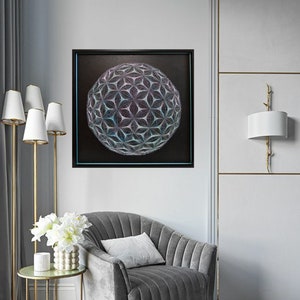 Op de afbeelding: Een grijze fluwelen fauteuil met een wit kussen staat in een kamer met een zwart ingelijst beeld van een geometrische bol met een blauwe en paarse gradiënt. Het beeld hangt aan een witte muur met gouden versiering. Naast de fauteuil staat een gouden bijzettafel met een witte vaas met witte bloemen.