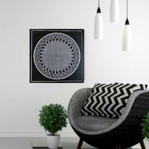 Op de afbeelding: Een zwart-wit mandala-ontwerp op een zwart canvas, hangend aan een witte muur boven een grijs-zwarte fauteuil met een zwart-wit gepatroneerd kussen. Drie witte hanglampen hangen aan het plafond.
