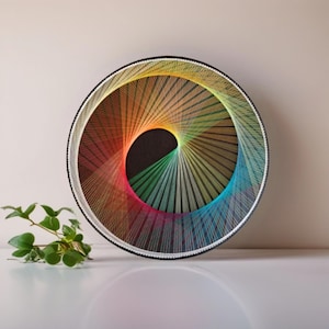 May include: A round string art piece with a black center and a rainbow of colors radiating outwards. The artwork is framed in a black circular frame with a white border. A small green plant is in the foreground.