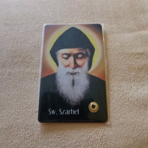 Könnte beinhalten: Eine religiöse Karte mit einem Bild eines Mannes mit Bart und schwarzem Gewand. Die Karte hat einen goldenen Heiligenschein um den Kopf des Mannes und den Text "Sw. Scharbel" unten.