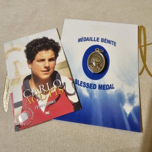 Könnte beinhalten: Eine silberne Medaille mit einem Bild eines jungen Mannes. Die Medaille liegt auf einer blauen Karte mit den Worten "MÉDAILLE BÉNITE" und "BLESSED MEDAL". Neben der Karte befindet sich ein Foto des jungen Mannes mit dem Text "CARLO ACUTIS (1991-2006)".