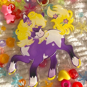 May include: A purple and white horse charm with a gold star charm attached. The horse has a yellow mane and tail with pink flowers. The charm is surrounded by colorful beads.