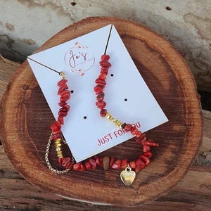 Op de afbeelding: Een rode koraal ketting- en armbandset met gouden accenten en een hartvormige bedel. De sieraden worden gepresenteerd op een houten oppervlak met een witte kaart met de tekst "Jo's" en "JUST FOR YOU".