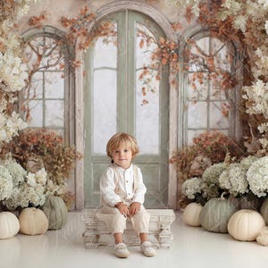 May include: A child wearing a white long-sleeved shirt and light beige trousers sits on a small white bench. The scene is framed by large arrangements of dried white and brown flowers, with several pale pumpkins placed on the floor.