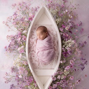 May include: A baby wrapped in a pink blanket lies in a white boat-shaped prop, surrounded by purple and white flowers. The background is a soft, pastel-coloured backdrop.