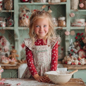 May include: A young girl in a floral apron and red long-sleeved shirt smiles at the camera. A bowl of flour and a wooden spoon sit on a wooden table. The background features a kitchen with shelves, gingerbread houses, and a red stand mixer.