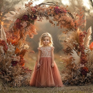 May include: A young girl in a peach-colored tulle dress stands in front of a floral archway decorated with dried flowers and pampas grass. The archway is made of branches and is adorned with fall-colored flowers.