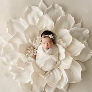 May include: A newborn baby sleeping in a white flower-shaped prop. The baby is wrapped in a white blanket and wearing a white headband with a flower.