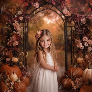 May include: A young girl wearing a white dress stands in front of a autumnal backdrop. The backdrop features a rustic archway decorated with autumn leaves and flowers, and pumpkins are scattered on the ground.