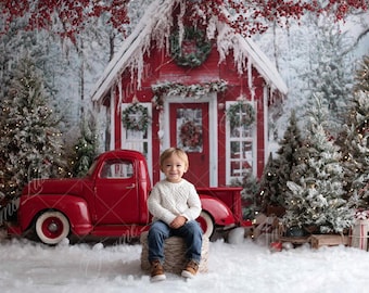 May include: A young child sits in front of a red vintage truck and a red cabin decorated for Christmas. The scene includes snow-covered evergreen trees, wreaths, and a snowy backdrop, creating a festive holiday setting.