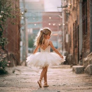 Könnte beinhalten: Ein junges Mädchen in einem weißen Tutu und Ballettschuhen steht in einer Gasse. Sie hat blondes Haar und trägt ein zartes Haaraccessoire. Der Hintergrund zeigt Backsteingebäude und sanfte Beleuchtung, was eine verträumte Atmosphäre schafft.