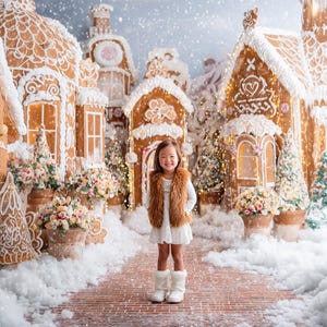 Puede incluir: Una niña pequeña se encuentra en una escena de un pueblo de pan de jengibre caprichoso. El fondo presenta casas de pan de jengibre detalladas con detalles de glaseado blanco, tejados cubiertos de nieve y coronas decorativas. La niña lleva un vestido blanco, un chaleco marrón y botas blancas.