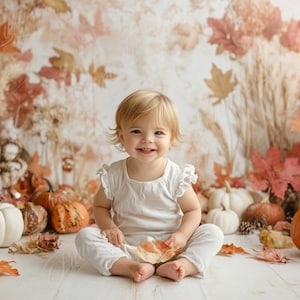 May include: A smiling baby girl wearing a white dress sits on a white wooden floor surrounded by fall-themed decorations, including pumpkins and leaves.