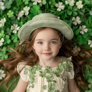 May include: A young girl with long brown hair wearing a green hat and a white dress with green clover accents lies on a bed of green shamrocks.