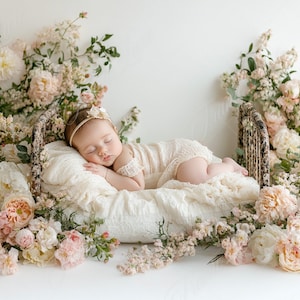 May include: A sleeping baby wearing a white lace dress and a headband lies in a wicker basket bed surrounded by pink and white flowers.