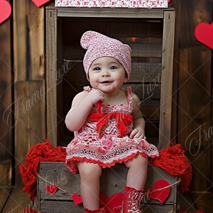 May include: A baby in a red and white dress and matching hat sits in a wooden box decorated with red hearts. The baby wears red patent leather boots and the sign above reads "kissing booth".