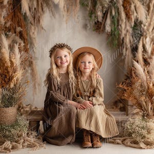 May include: Two young girls in matching brown dresses and floral crowns sit together, smiling. One wears a hat. The background features dried plants and a pumpkin, creating a warm, autumnal scene. The girls are sitting on a wooden bench.