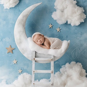 Op de afbeelding: Een pasgeboren baby slaapt op een witte halve maan prop, omringd door witte wolken en gouden sterren op een blauwe achtergrond. De maan staat op een witte houten ladder.
