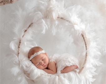May include: A newborn baby sleeping in a white feather-filled hoop, suspended from a branch. The baby is wearing a white lace dress and a white headband.