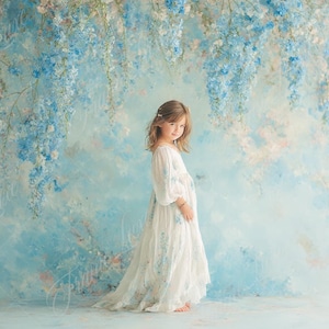 May include: A young girl in a white dress with blue floral accents stands before a backdrop of blue and white flowers. The dress has long sleeves and a flowing skirt. The background is a painted mural with blue and white wisteria.