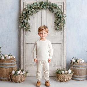 Op de afbeelding: Een jonge jongen in een crèmekleurige trui en broek staat voor een rustieke houten deur die versierd is met een krans van groen. De jongen glimlacht en kijkt naar de camera. Aan weerszijden van de deur staan ​​houten vaten met decoratieve eieren en groen.