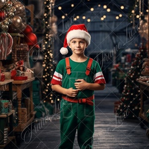 May include: A young person wearing a green elf costume and a red Santa hat stands in front of a Christmas-themed backdrop. The backdrop features a wooden shelf with Christmas decorations, a Christmas tree, and a red pillow.