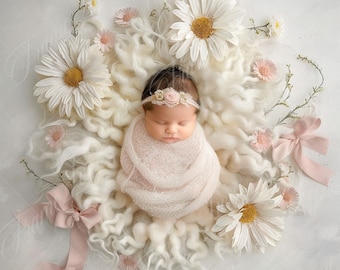 May include: A newborn baby girl is sleeping peacefully in a white fluffy nest surrounded by white and pink daisies. She is wearing a pink swaddle and a flower headband.