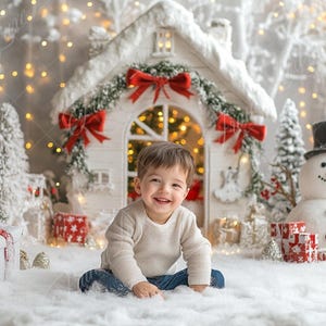 May include: A young child sits in a snowy winter wonderland setting. The child is wearing a white sweater and blue jeans. There is a white toy house with red bows and a snowman in the background.