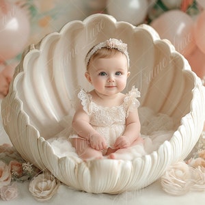 Peut inclure: Une petite fille vêtue d'une robe en dentelle blanche et d'un bandeau blanc avec une fleur est assise dans un accessoire en forme de coquille blanche. L'arrière-plan est décoré de ballons et de fleurs roses et blancs.