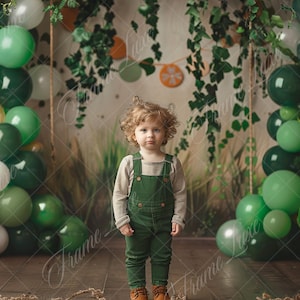 May include: A young child with curly hair wearing a green overall and a light gray long-sleeved shirt stands in front of a green and white balloon archway. The background is a green and brown backdrop with vines and leaves.