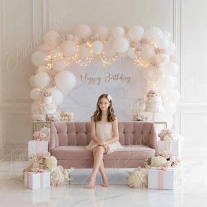 Op de afbeelding: Een jong persoon zit op een roze, gecapitonneerde bank voor een achtergrond met ballonnen, bloemen en een "Happy Birthday" banner. De scène bevat twee taarten, cadeaus en lichtslingers, wat een feestelijke sfeer creëert.