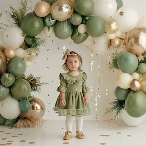 Puede incluir: Una niña pequeña con un vestido verde con cuello de volantes se encuentra frente a un arco de globos verde, dorado y blanco. Los globos están dispuestos en forma de semicírculo y están decorados con purpurina dorada.