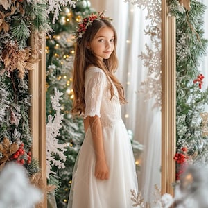 May include: A young girl wearing a white lace dress and a red berry wreath in her hair stands in front of a mirror decorated with snowflakes and greenery. The mirror is framed in gold and surrounded by a wintery scene.