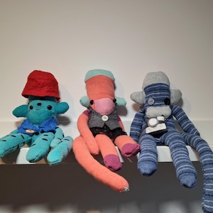 May include: Three handmade sock creatures. The first is teal with a red bucket hat and blue jacket. The second is coral with a grey waistcoat and black shorts. The third is blue striped with a grey hat and camera.