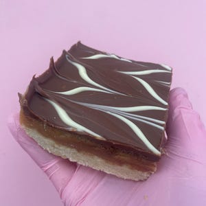 Könnte beinhalten: Ein quadratischer Dessertriegel mit einem Shortbread-Boden, einer Karamellschicht und einem Schokoladenbelag mit weißen Wirbeln. Das Dessert wird von einer behandschuhten Hand vor einem rosa Hintergrund gehalten.