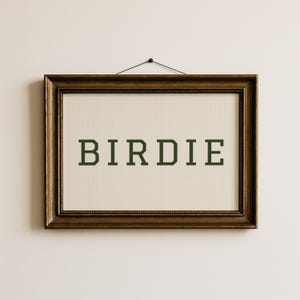 Puede incluir: Una obra de arte en punto de cruz enmarcada con la palabra "BIRDIE" en letras de bloque verde oscuro. La obra de arte est&aacute; sobre un fondo color crema y enmarcada en un marco de madera marr&oacute;n oscuro y ornamentado. El marco est&aacute; colgado en una pared blanca.
