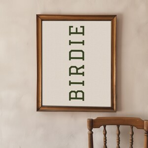 Puede incluir: Una obra de arte enmarcada con la palabra "BIRDIE" en verde oscuro, centrada verticalmente sobre un fondo crema cuadriculado. El marco es de madera oscura. La obra est&aacute; colgada en una pared beige claro.