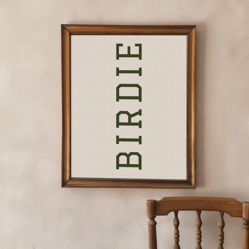 Puede incluir: Una obra de arte enmarcada con la palabra "BIRDIE" en verde oscuro, centrada verticalmente sobre un fondo crema cuadriculado. El marco es de madera oscura. La obra est&aacute; colgada en una pared beige claro.
