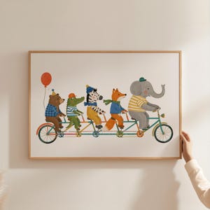 Puede incluir: Una impresión enmarcada con una ilustración caprichosa de cinco animales antropomórficos montando en bicicleta tándem. Los animales incluyen un oso, un caimán, una cebra, un zorro y un elefante, cada uno con ropa. Un globo rojo flota sobre el oso.