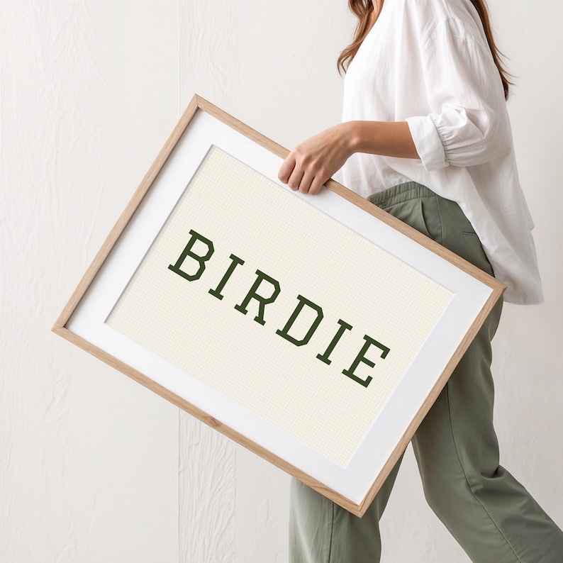 Puede incluir: Una obra de arte enmarcada con la palabra "BIRDIE" en letras may&uacute;sculas de color verde oscuro. La obra tiene un fondo color crema y est&aacute; enmarcada en un marco de madera clara con un passe-partout blanco. El marco lo sostiene una persona.
