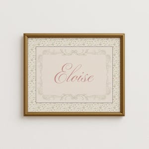 May include: A framed print featuring the name "Eloise" in a cursive script. The print has a pale pink background, a decorative border, and a gold frame. The border is adorned with a floral design.