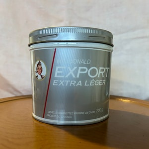 May include: A silver can of McDonald Export Extra Léger tobacco. The can has a red stripe with the brand name and a picture of a woman in a red dress. The can is labeled "Tabac à cigarettes Virginie de choix 200g Produit du Canada".