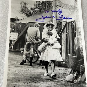May include: Black and white photograph of Jamie Farr in a costume, signed "To Marty, Jamie Farr." Farr is wearing a dress, hat, and boa, walking on a dirt path. A man on a bicycle is in the background.