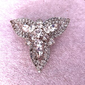 May include: A silver-toned brooch with a three-part design, heavily embellished with clear, sparkling rhinestones. The brooch features a central, elongated teardrop shape, flanked by two symmetrical, leaf-like sections. The rhinestones vary in size and shape.