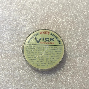 May include: A vintage Vicks VapoRub tin with a white and green label. The label reads "Improved White Stainless Vicks Vapour Rub" and includes text describing its uses. The tin is circular and has a metallic appearance.
