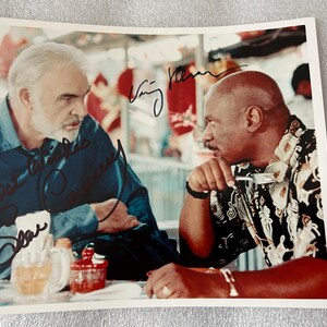 May include: A photograph featuring two men seated at a table, with autographs in black ink. One man wears a blue shirt, the other a patterned shirt. Drinks and condiments are on the table. The background is blurred.