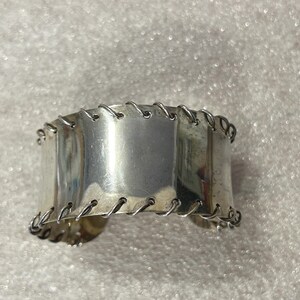May include: A wide, silver-toned cuff bracelet. The bracelet is made of rectangular metal plates connected by a series of small, curved metal links. The cuff has a polished, reflective surface and a simple, industrial design.