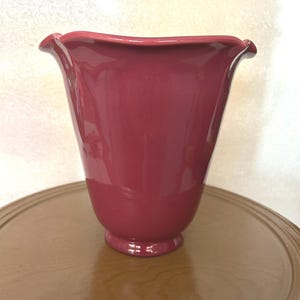 May include: A glossy, burgundy-colored vase with a flared, ruffled rim. The vase has a rounded body and a small, circular base. The vase is sitting on a wooden surface.