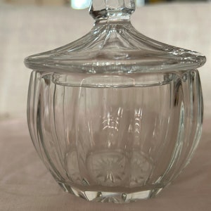 May include: A clear glass jar with a lid. The jar has a decorative pattern on the bottom and sides. The lid is domed and has a knob on top.