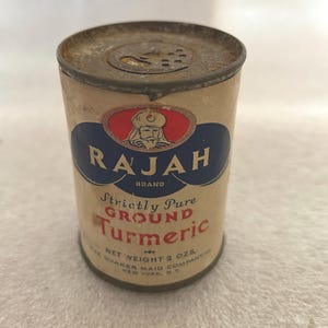 May include: A vintage metal tin of Rajah brand Strictly Pure Ground Turmeric. The tin is white with blue and red accents and features a drawing of a man in a turban. The text on the tin reads "Rajah Brand Strictly Pure Ground Turmeric Net Weight 2 ozs The Quaker Maid Company New York, NY."